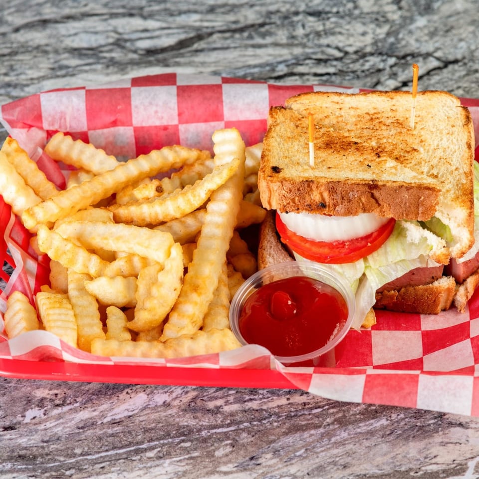 A fried bologna sandwich with lettuce, tomato, and onion, served with crinkle-cut fries and ketchup.